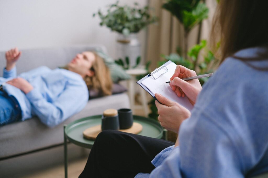 A therapy session in progress with a therapist taking notes as a patient reclines on a sofa.